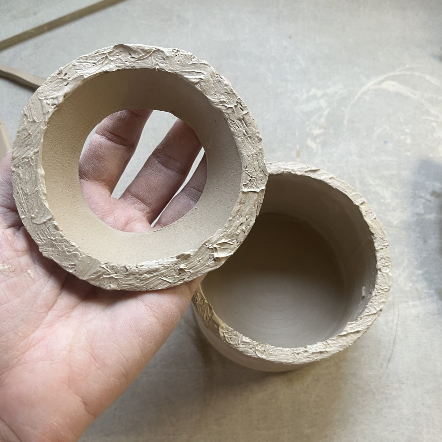Close-up of two handmade clay pieces being prepared for joining to for the body of the teapot. The edges are scored and coated with slip, showcasing the hand-building process in pottery making. 