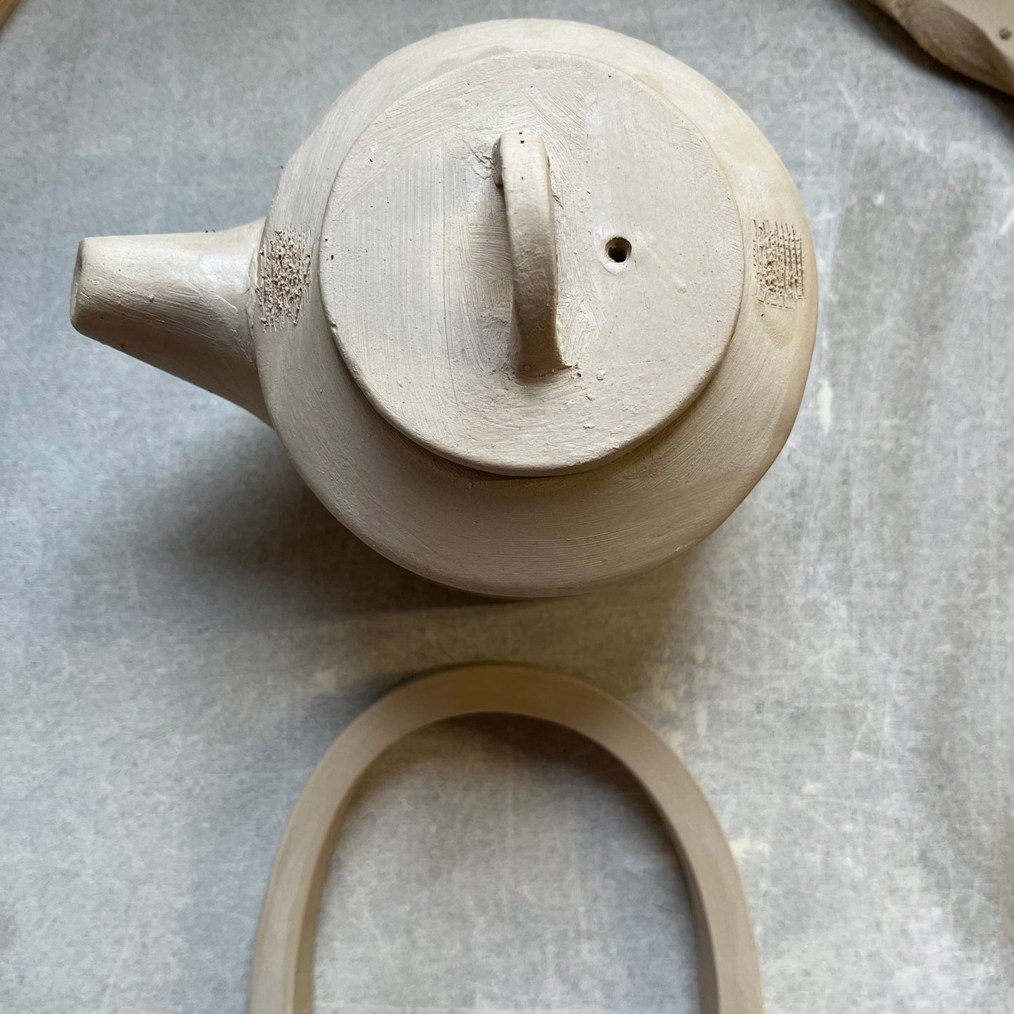 Top view of a handmade ceramic teapot with a fitted lid featuring a small handle and steam hole. A curved clay handle lies below, ready to be attached, showcasing the hand-building process in pottery. Apart of a Printable Pottery Teapot for sale .