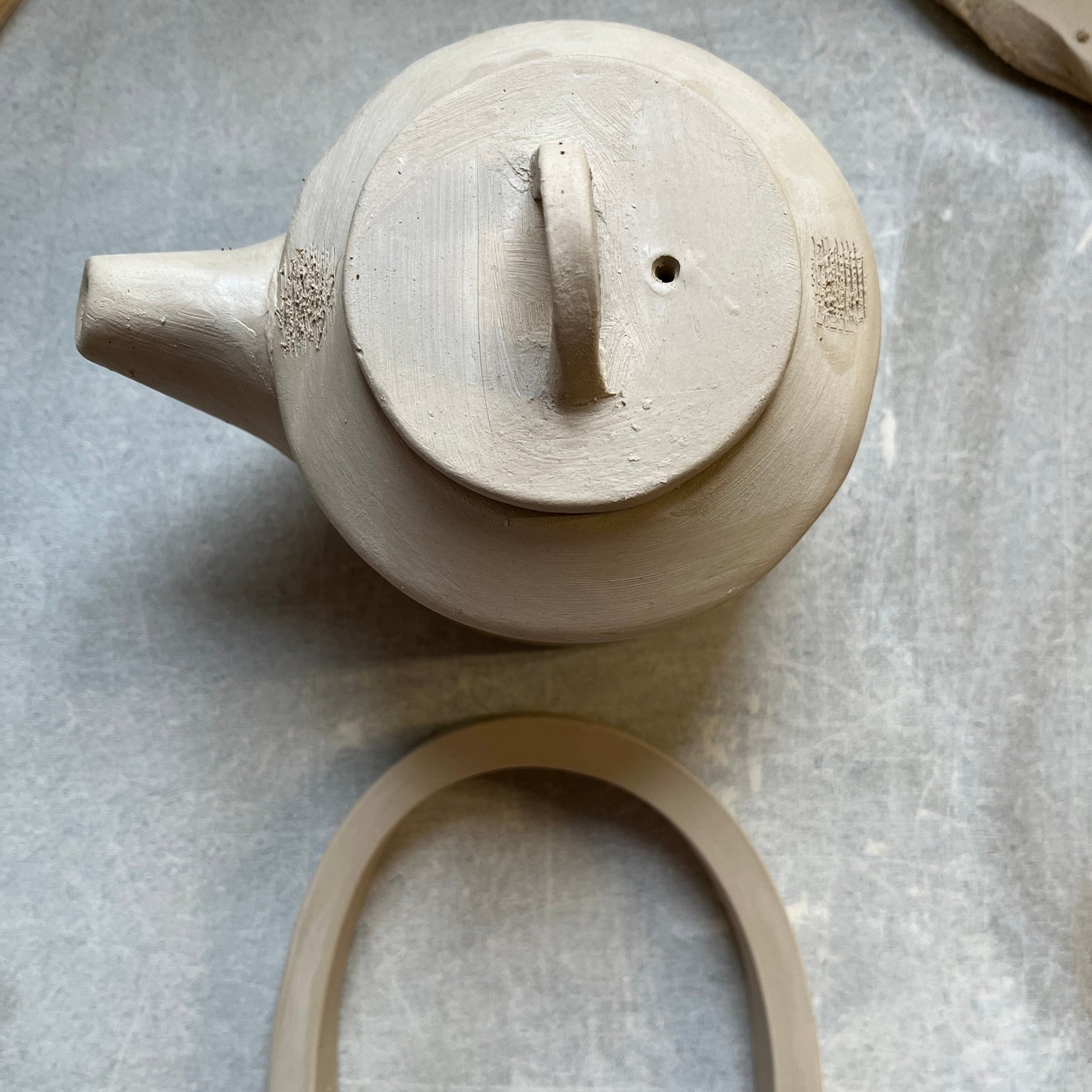 Top view of a handmade ceramic teapot with a fitted lid featuring a small handle and steam hole. A curved clay handle lies below, ready to be attached, showcasing the hand-building process in pottery. Apart of a Printable Pottery Teapot for sale .