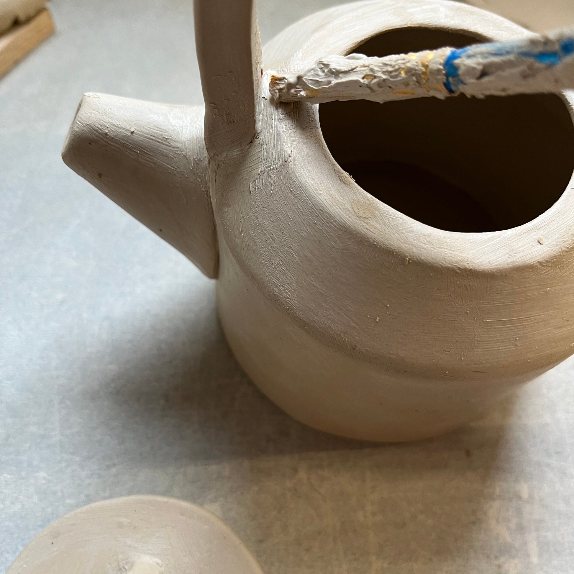 Close-p of a handmade ceramic teapot in progress with a brush applying slip to attach a curved handle to the body. The lid rests nearby, showcasing the detailed pottery hand- building process 